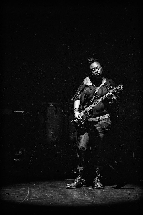 A black and white photo of Tamar Kali. She wears a long off-the-shoulder sweater and tights. She closes her eyes as she plays her guitar onstage.
